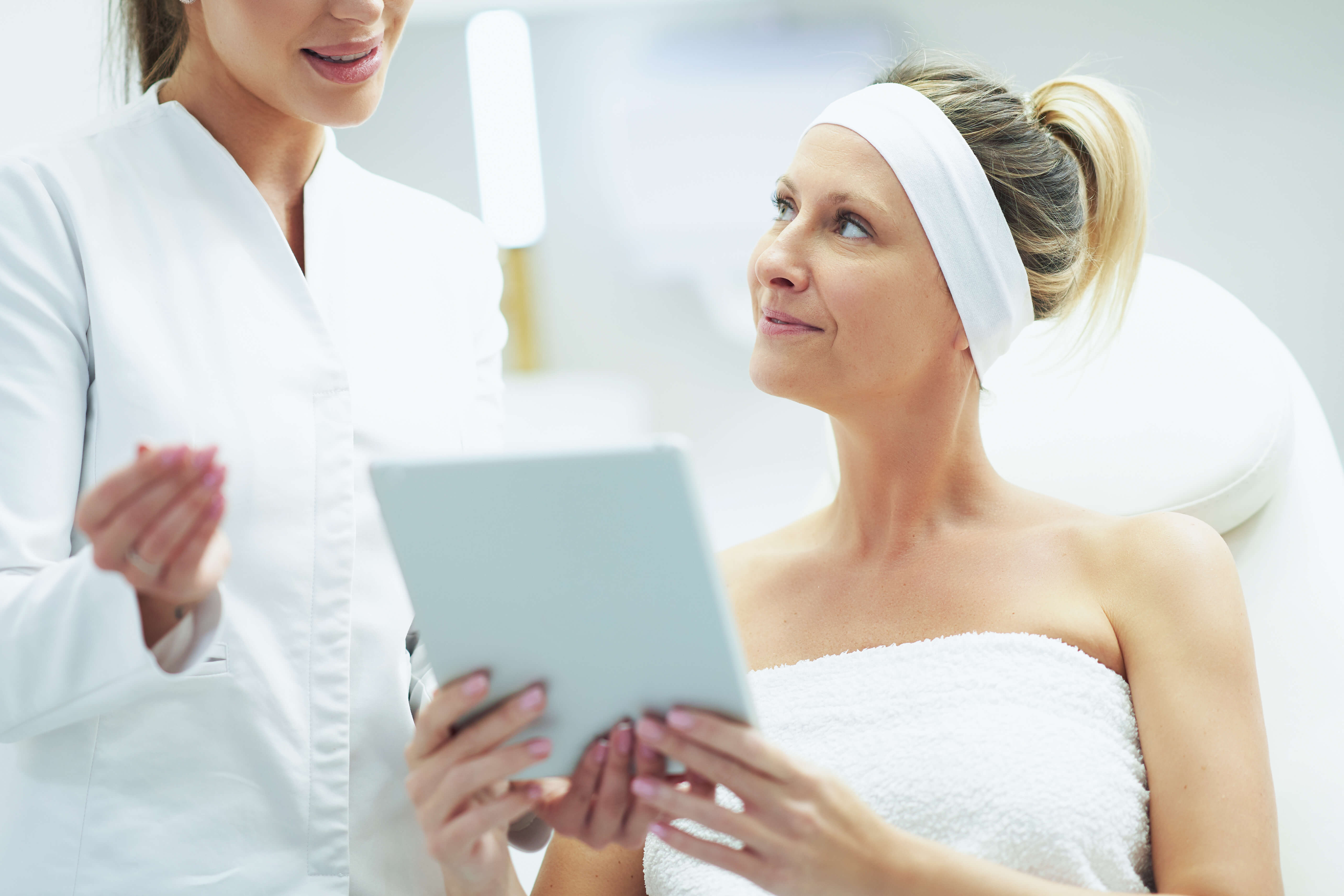 Woman in a spa setting holding a tablet while another woman assists her.