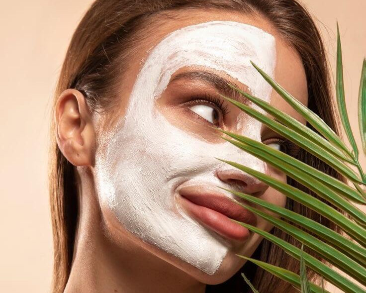 Woman with a white facial mask holding a green leaf against her face on a beige background